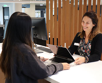 A friendly Library staff member issues a laptop to a young student at the Library enquiry desk at Joondalup Campus.