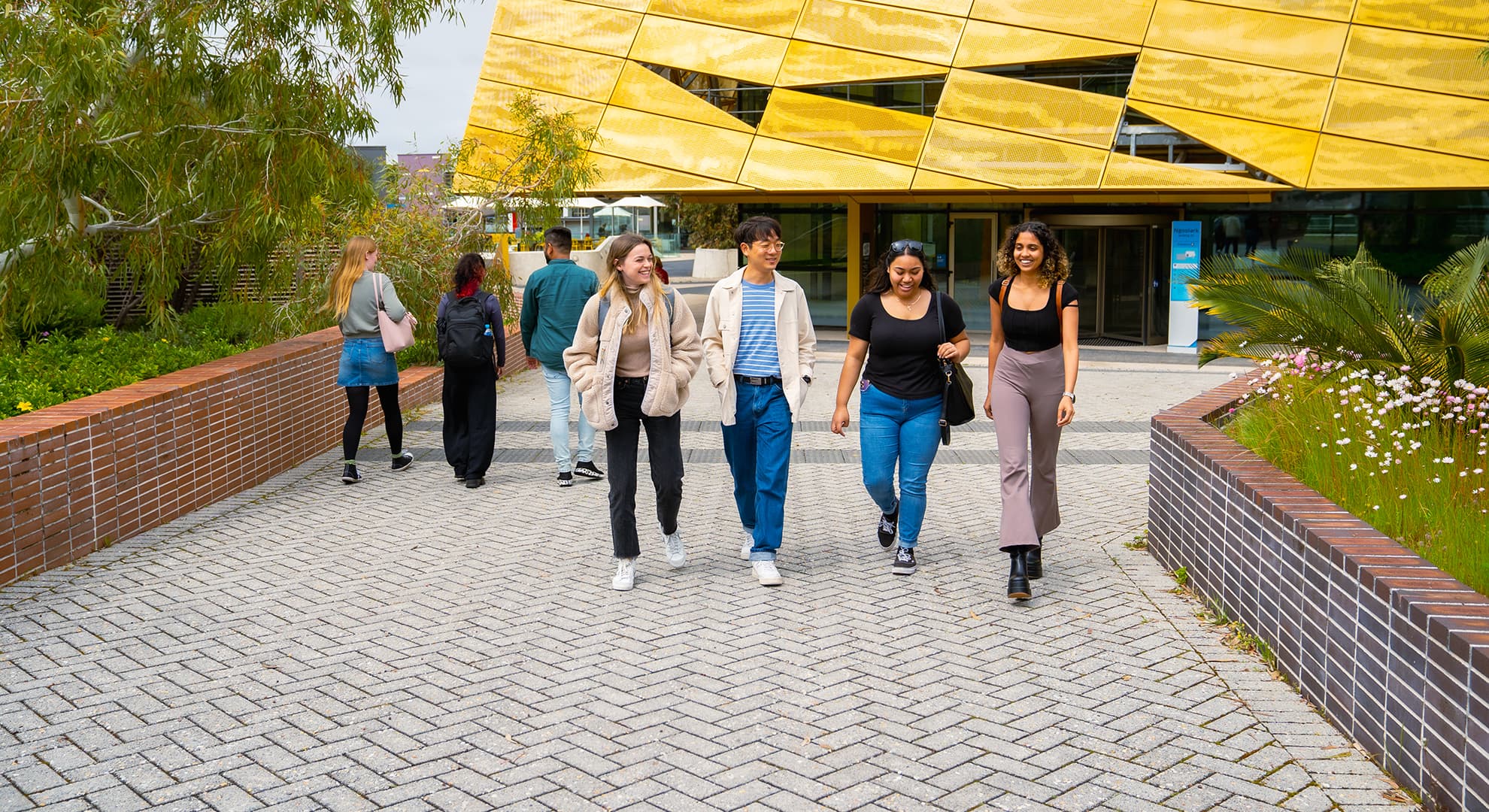 A group of Edith Cowan University students walking around Joondalup Campus. There is building in the background with gold geometric panelling.