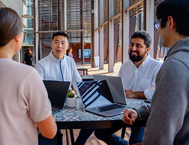 Four people sit at a cafe on a university campus with their laptops, having a discussion.