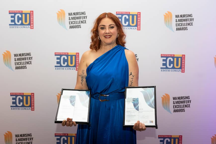 Sandra Vinciguerra is wearing a blue one-shoulder gown and holds out her two awards and smiles.