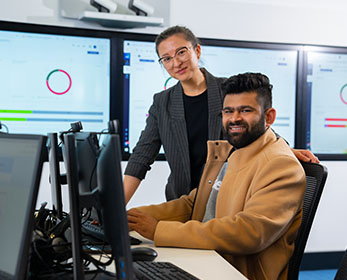 A smiling student sits at a computer in a learning environment setting, with another person assisting them with their work.