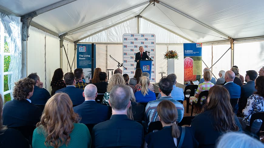 View into the marquee of VC addressing guests at lectern with 'The Living Room' banner on the left of the stage