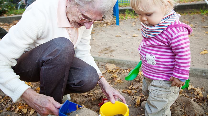 Elderly white haired lady with toddler squatting playing in a sand pit