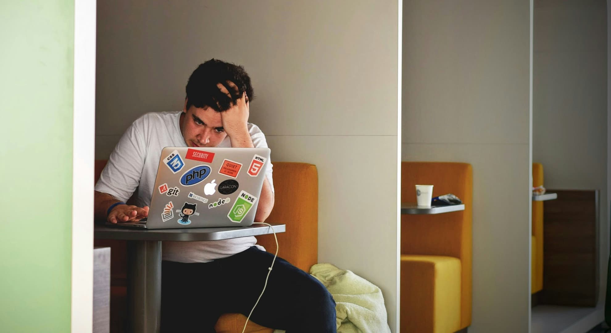 Young man sitting at desk with laptop with stickers on it with his head in his hand