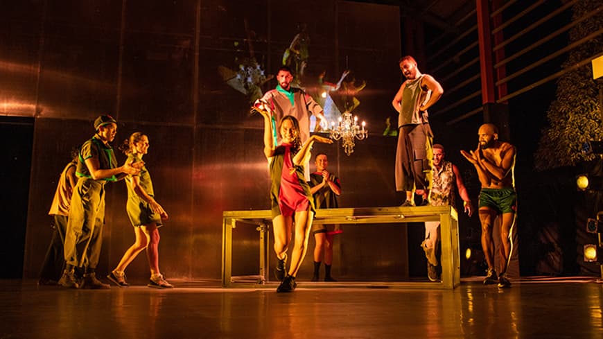 A theatre performance on stage, a woman angles her arms in movement while other people watch on in encouragement.