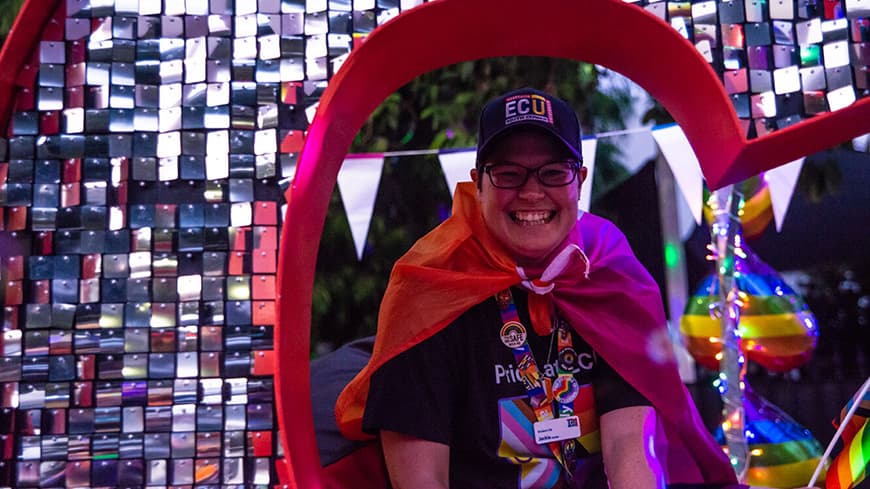 Person in ECU branded t-shirt, cap and lanyard, with red cape tied around neck sitting in the crook of a giant letter \'C\' made from red metal and covered in little silver squares of shiny tinsel