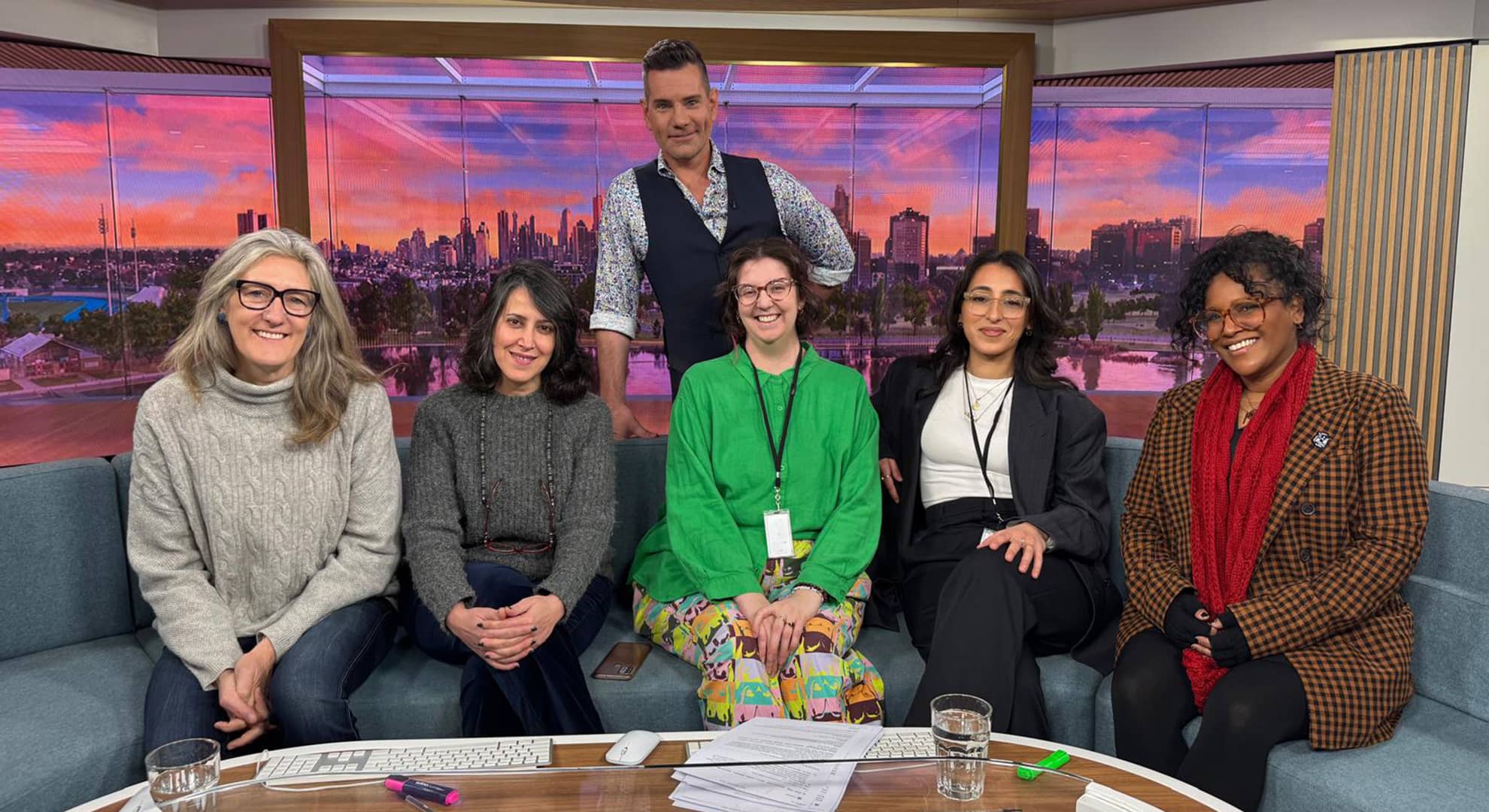 Five people sitting on a TV studio couch facing the camera with the host standing behind them