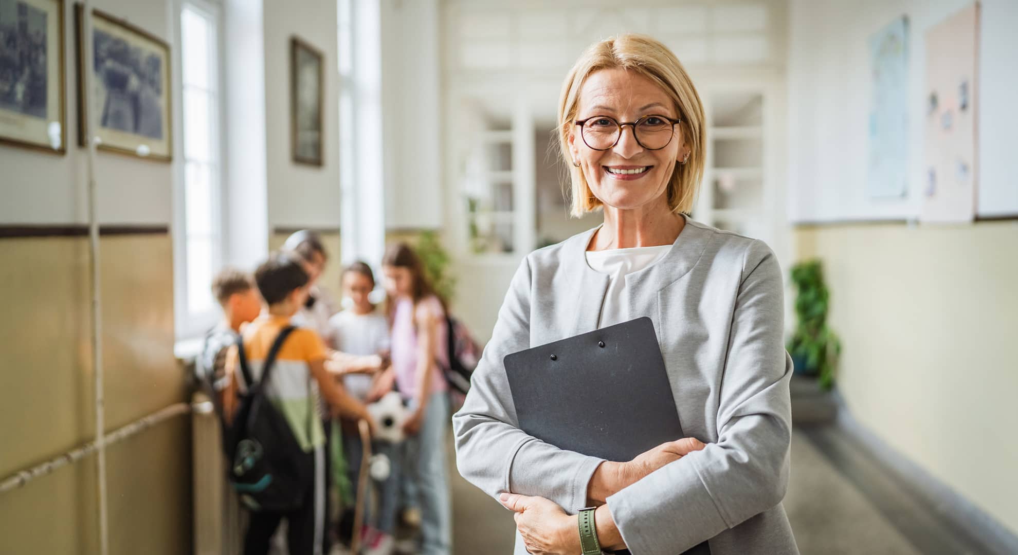 Portrait of mature woman teacher standing in school hallway with clipboard, smiling at camera. She has black framed glasses and short blonde hair. A group of children are blurred in the background interacting with one another.