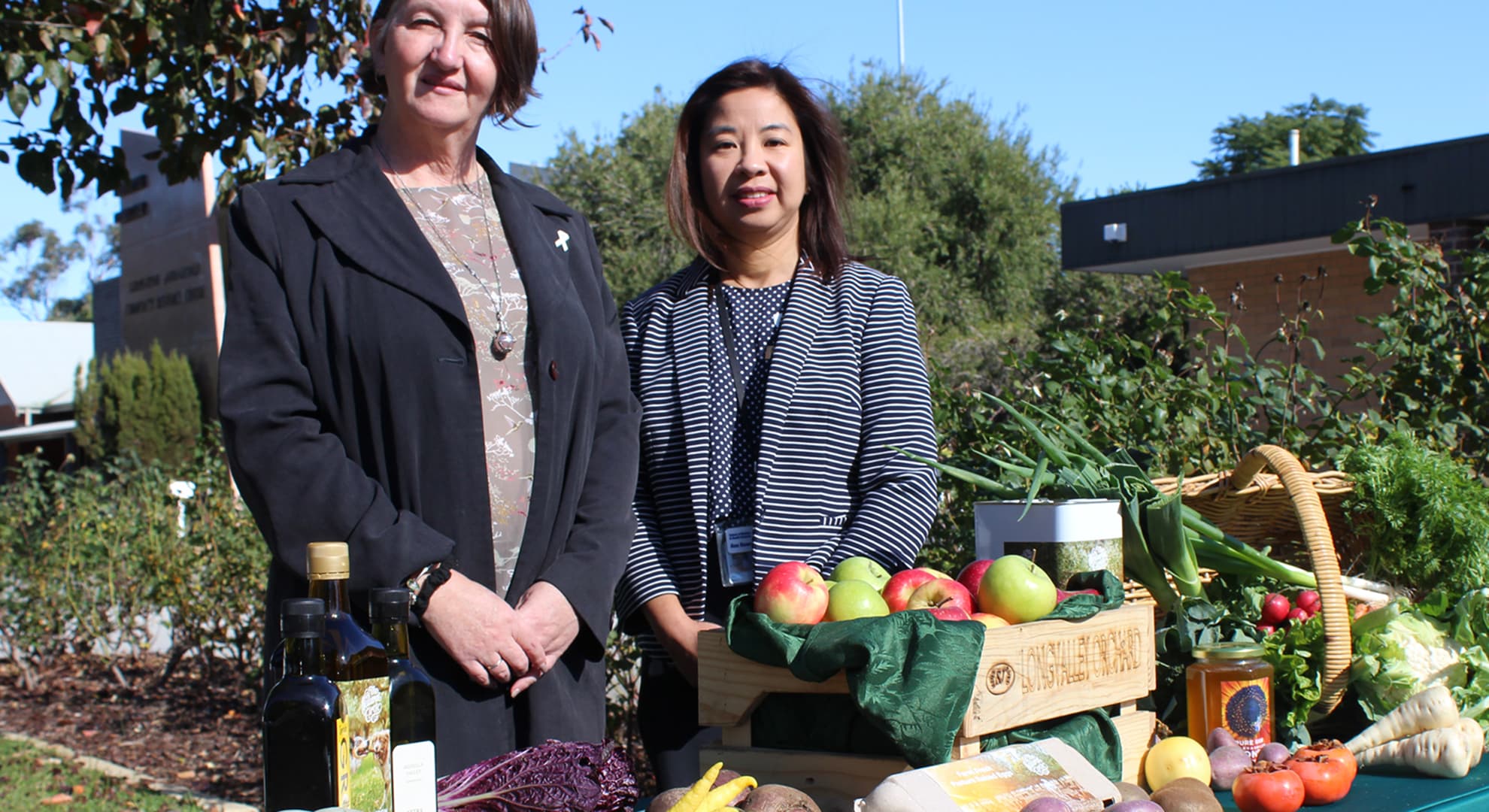 Two women stand behind table full of fresh fruit and vegetables