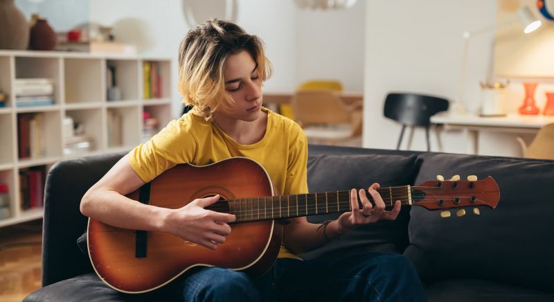 Teenage boy sitting on a couch in a home with a yellow shirt looking down and playing guitar.