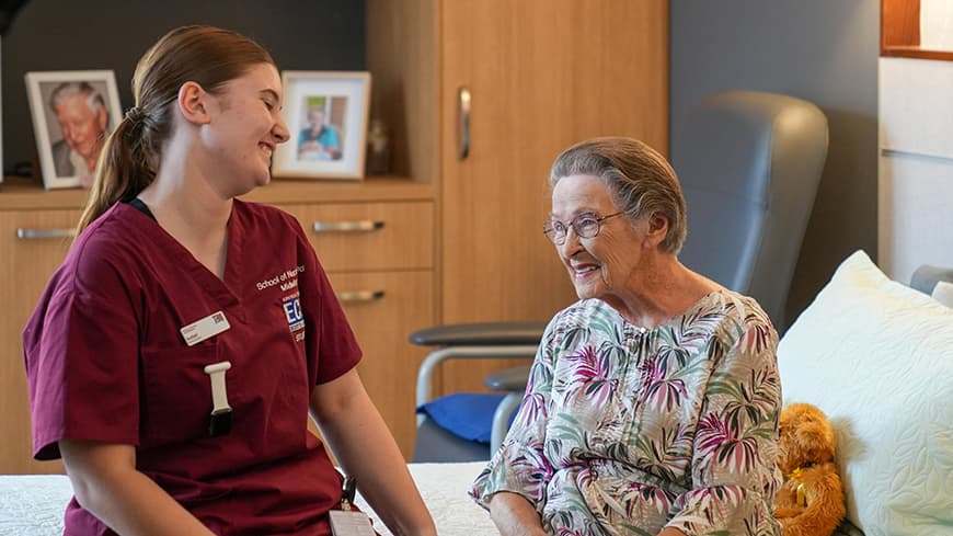 A young nursing student and an aged care resident sit on a bed in the middle of a joyful conversation.