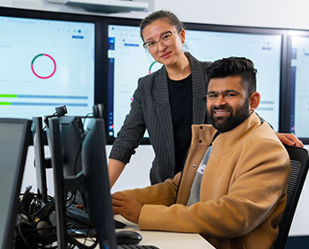 A student in a computer lab setting smiles down the lens of a camera, and a teacher stands behind him.