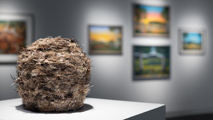 Foreground round feather vessel on white gallery display plinth with blurred paintings on walls in background