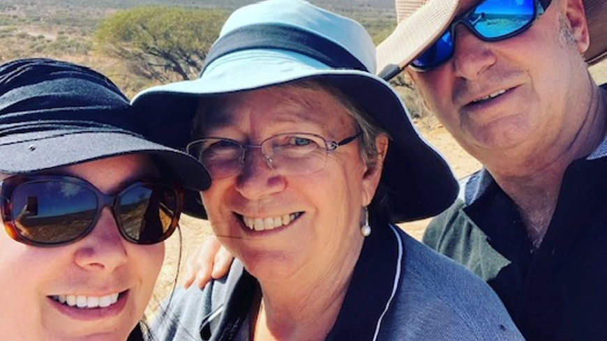 Three people close up, L-R young woman in cap and sunglasses, older woman in glasses and a blue hat and older man in light hat and sunglasses with Australian desert in background