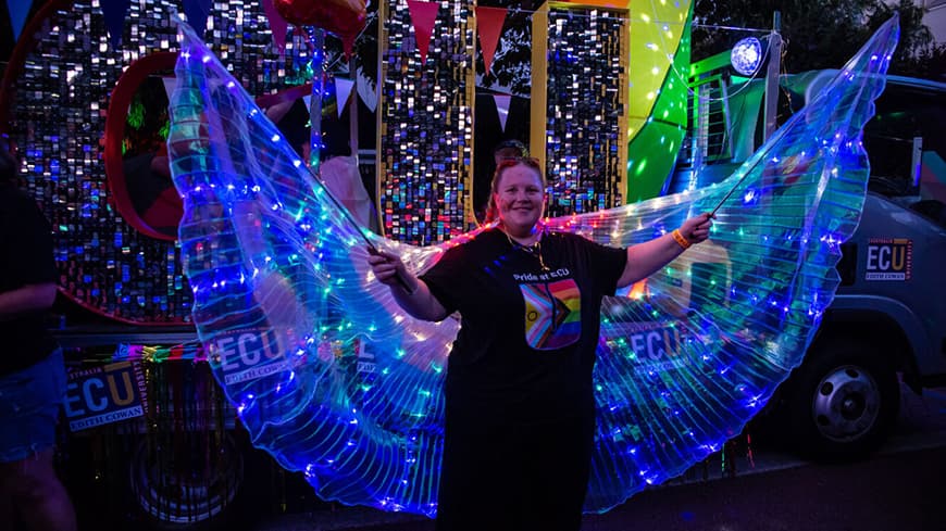 Person in black pants and an ECU branded Pride t-shirt holding out lit-up sparkly wings to their side, standing in front of ECU's Pride float truck with the giant letters ECU
