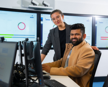 Two students in a computer lab, one seated at a workstation and the other standing beside them, both smiling. Large screens in the background display data visualizations.