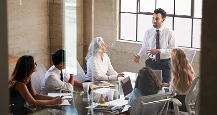 Four people sitting down at a boardroom table, looking up at man presenting