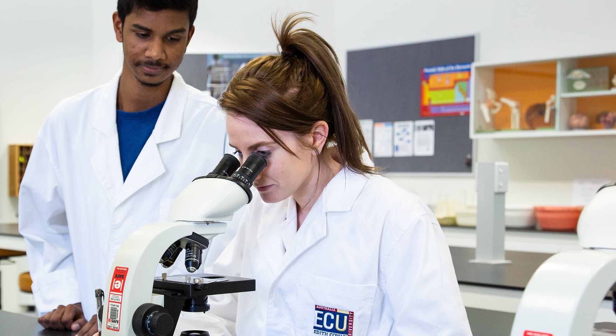 Two students in lab coats, one looking into a microscope.
