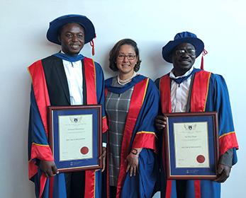 Photo of 2 male PhD graduates in regalia with their supervisor standing in the middle