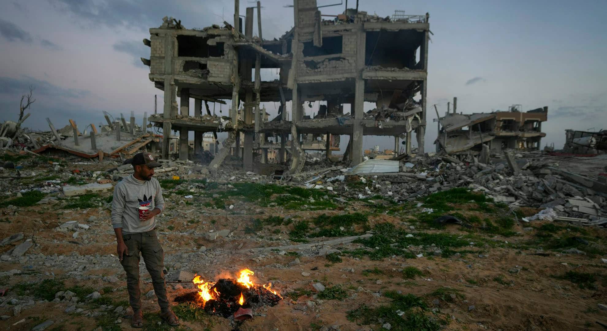 Man standing in front of a fire, with a demolished building in the background.