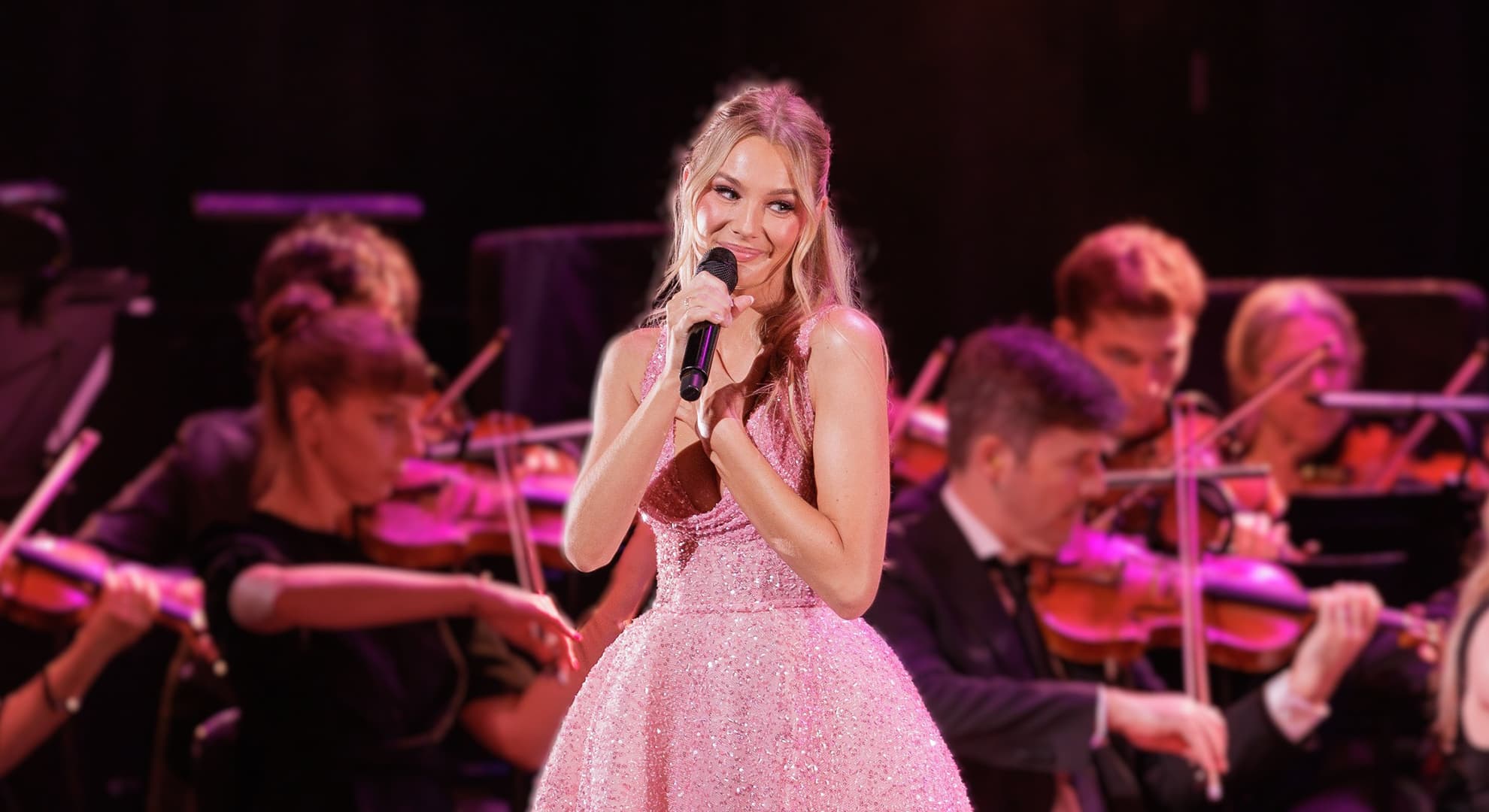 Amy Manford performing on stage in a Pallas Couture 'Aurora' pink ball gown. A live orchestra can be seen positioned behind her.