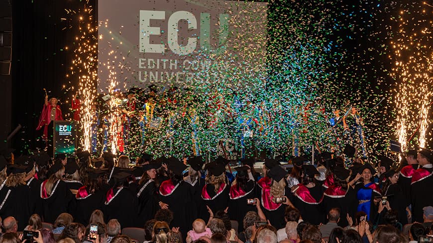 Graduates celebrate on and in front of a stage as streamers and indoor fireworks go off
