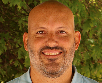 Headshot image of a smiling bald-headed man in a blue polo shirt