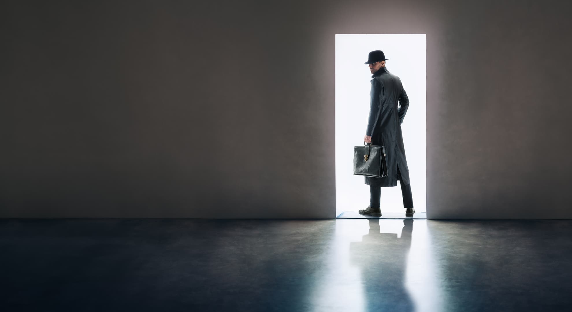 Man silhouette in hat and raincoat standing in the light of opening door in dark room