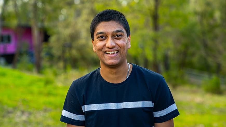 Kavan Patel has short black hair, wears a navy t-shirt with stripes and is outdoors