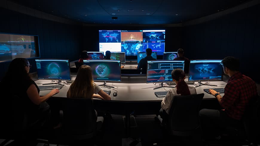 Eight people sit at computers in a cybersecurity operations centre. The computer monitors show maps of the world with graphs and data.