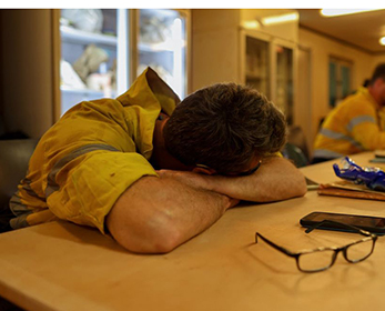 Image of a man with his head sleeping on a desk at work, wearing FIFO uniform