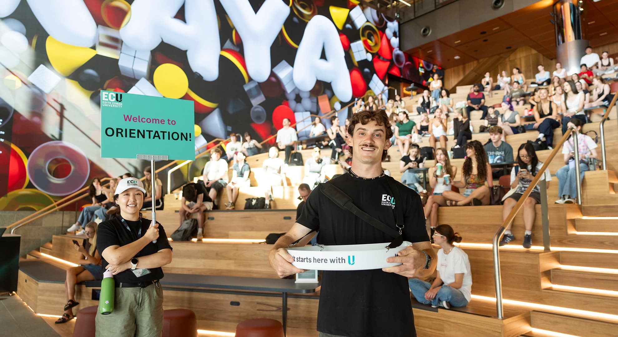 Two students standing in front of the ECU City Campus internal screen with banners