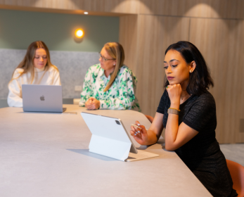 Three students seated at a study table in a modern library space using laptops and a tablet for study and research.