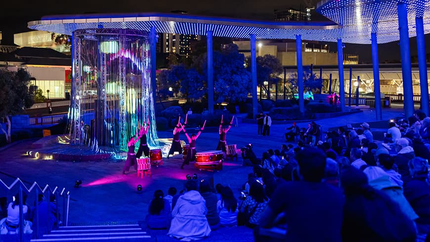 Japanese drummers standing in front of a crowd at night, backlit by string lights.