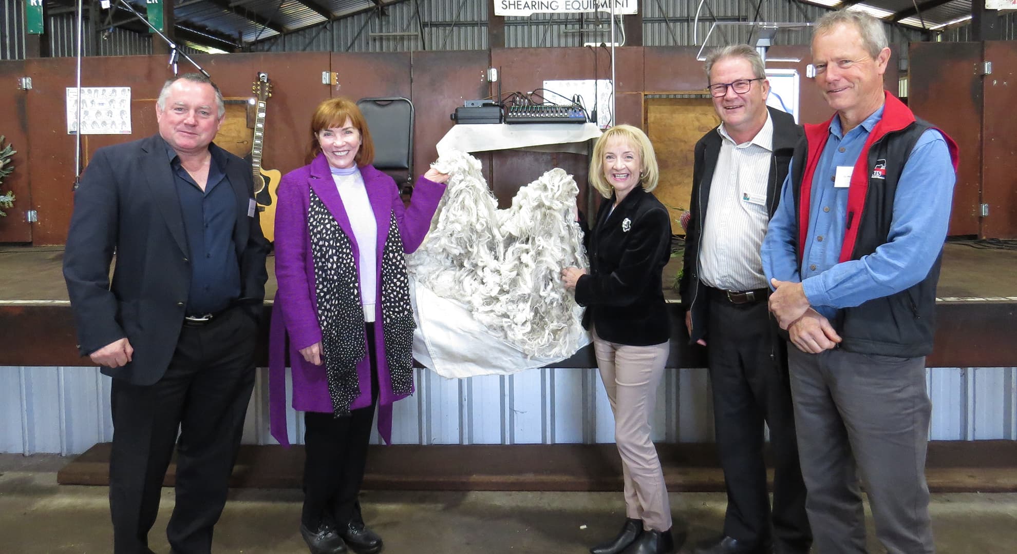 Five people standing in a shearing shed with two women holding the fleece in front of shearing equipment.