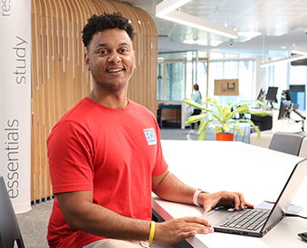 A student in a red t-shirt sits in the Library transitional space with a laptop, smiling at the camera. Need help with the technology you're using? Head to your Campus Library to find peer support. This service is also available online.