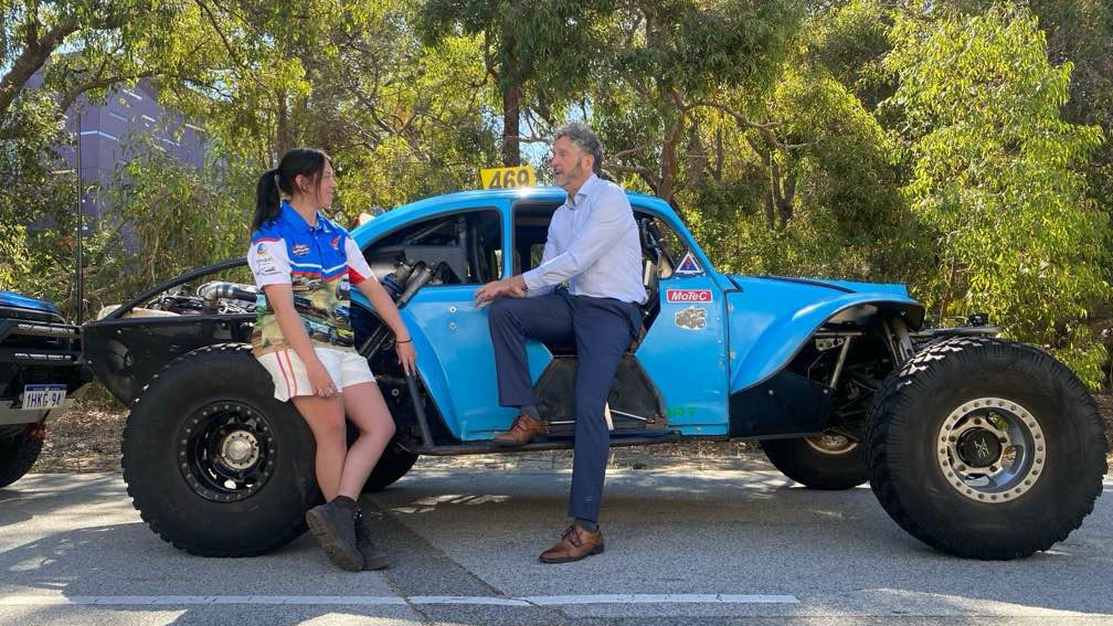 Woman on left in racing shirt and shorts leaning on blue racing car talking to man in navy pants and blue long sleeved shirt also leaning on car on the right