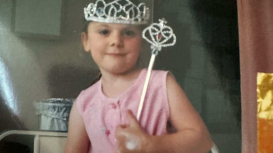 Little girl in pink dress, with plastic silver crown and wand sitting on a hospital bed with a hospital band on her wrist and a bandaid on her hand