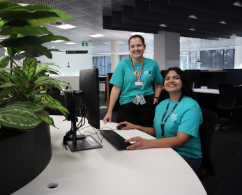 Two Library Peer Assistants wearing teal ECU shirts assisting students at a service desk in the ECU Library.