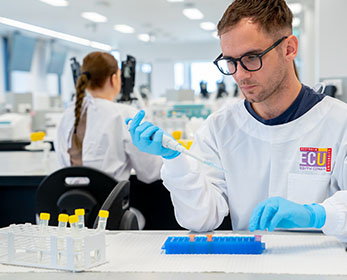 A student in an ECU branded white coat and safety googles is testing vials of liquid in a lab setting.