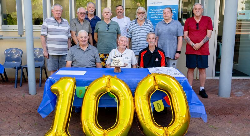 Kevin Ryan and his fellow Department of Veterans' Affairs group pose with the birthday cake, there are gold balloons shaped as the number 100 sitting in front of the table.