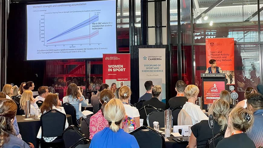 Professor Sophia Nimphius stands at the lectern delivering a presentation on women’s strength and conditioning to a full crowd.