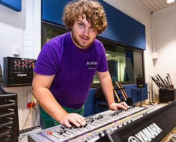 A young student commands use of a large mixing desk in a spacious recording studio full of musical equipment.