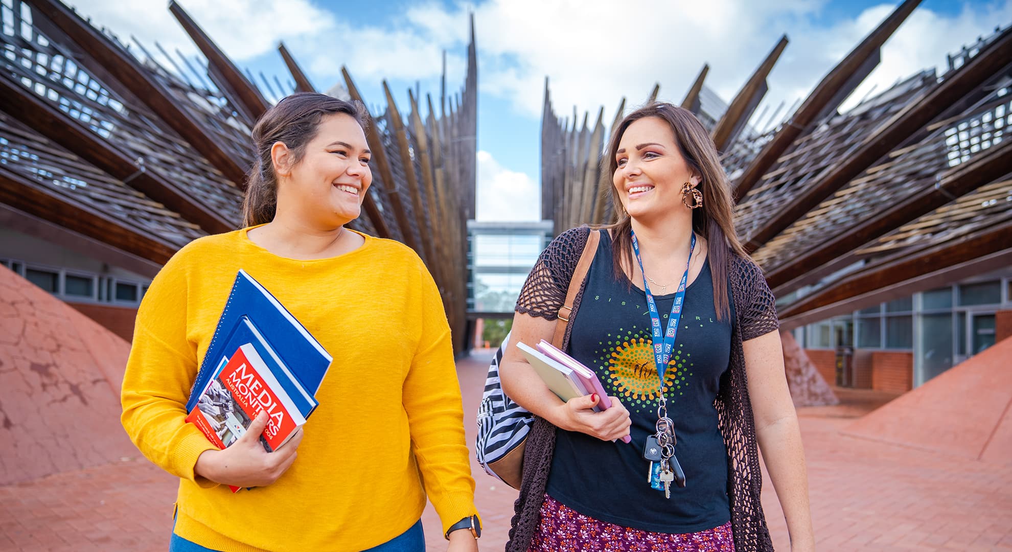 Two Indigenous students walk with textbooks in their hands smiling on the ECU Joondalup Campus.