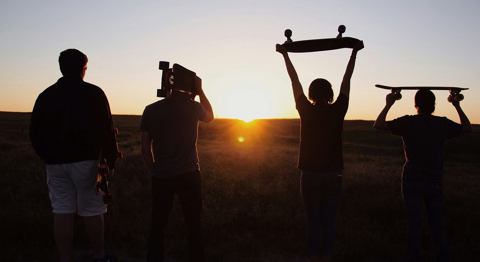Four people holding skateboards, two on the right holding them above their heads silhouetted against the sunset