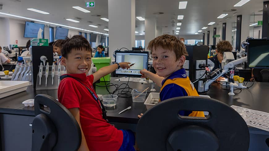 Two young students pointing at a computer monitor whilst smiling for the camera