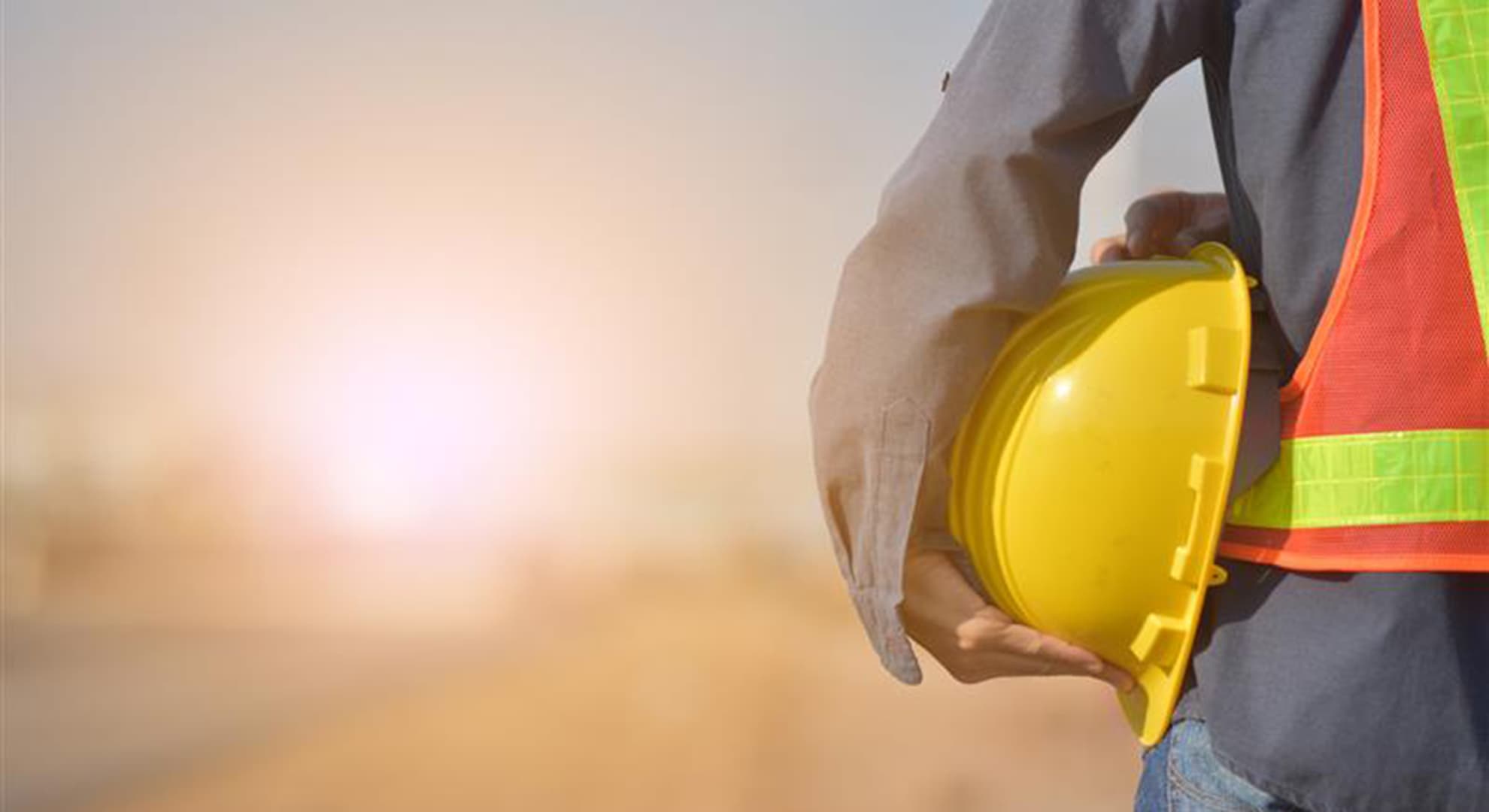 Man holding a hard hat facing a sunrise.