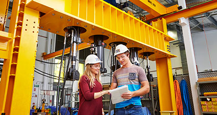 Woman and man wearing hard hats, standing in front of large engineering equipment.
