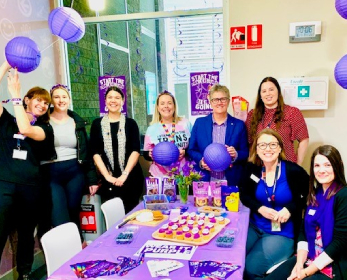 Eight staff members from ECU's Student Life team standing in front of a table of purple baked goods and holding purple paper lanterns.
