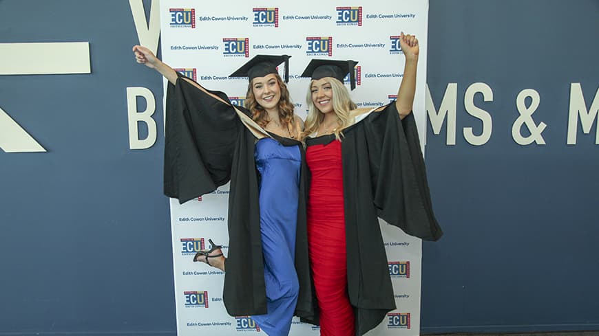 Left to right: sisters Lucy Norris and Hannah Norris celebrating their ECU graduation, wearing graduate regalia, holding each other with arms in the air celebrating.